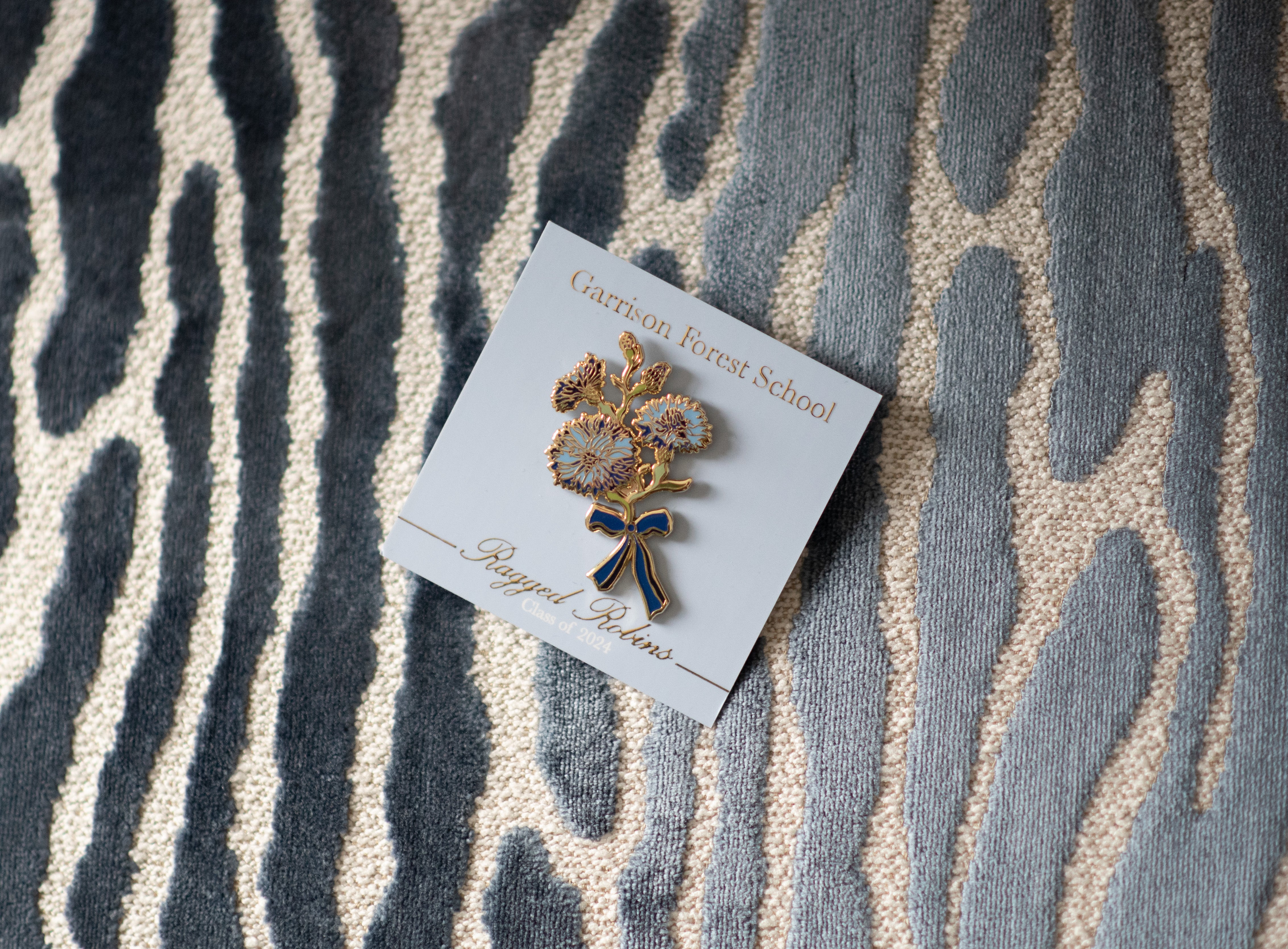 Decorative brooch on a card for the high school 'Garrison Forest School' text, placed on a textured surface.