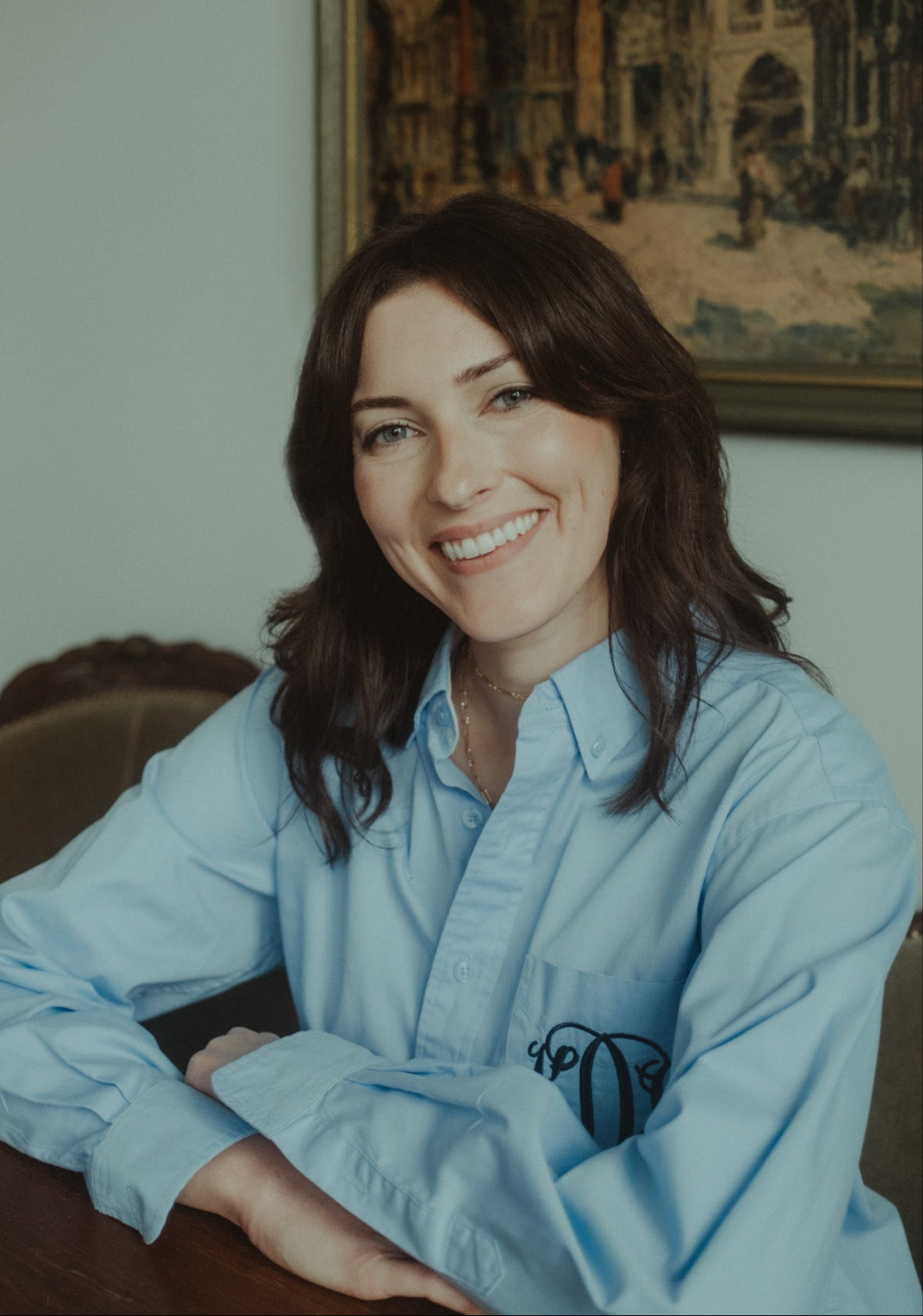 Founder of Doheny NYC Kate Doheny wearing a light blue shirt with a logo, sitting indoors with a painting in the background.